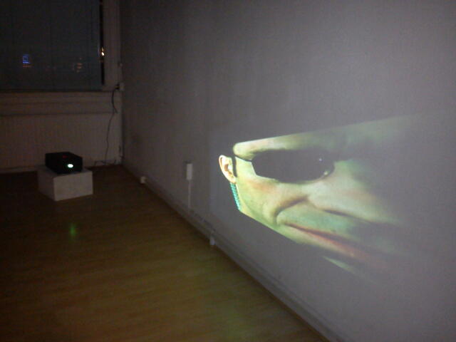 Projectie van agent Smith met zonnebril op de galeriemuur, de projector op een sokkel op de vloer